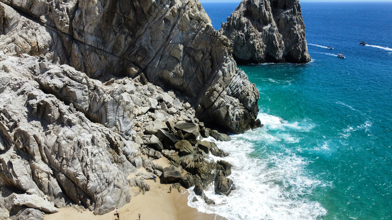 Rugged coastal cliffs meet the turquoise waters of the Pacific Ocean at Cabo San Lucas, Mexico. Waves crash against the rocky shoreline, while a small sandy cove nestled between the towering rock formations offers a secluded retreat. A few people explore the beach, and boats cruise in the distance, adding to the adventurous, scenic atmosphere.