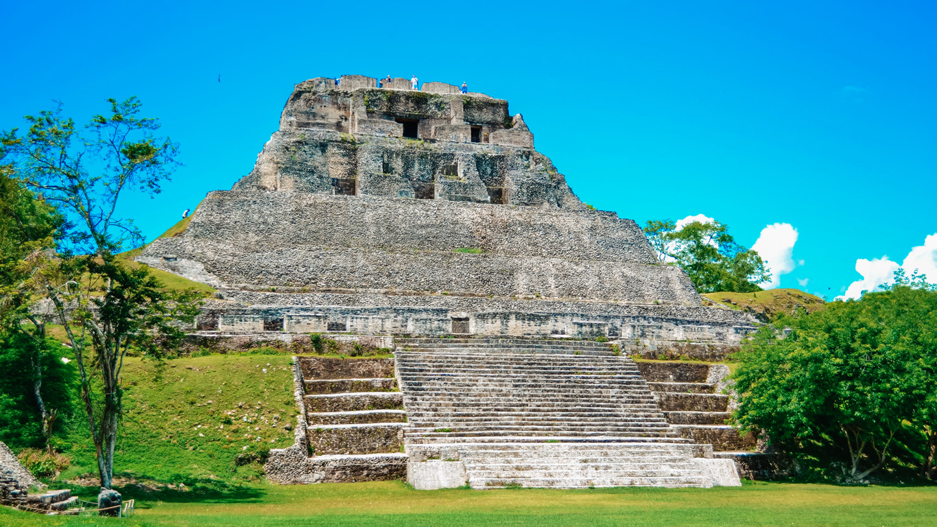 The ancient Mayan ruins of Xunantunich in Belize, featuring a grand stone pyramid with multiple levels and staircases. A few visitors stand at the top, taking in the panoramic view, while lush green trees surround the site. The bright blue sky and scattered clouds highlight the historic and cultural significance of this archaeological wonder.