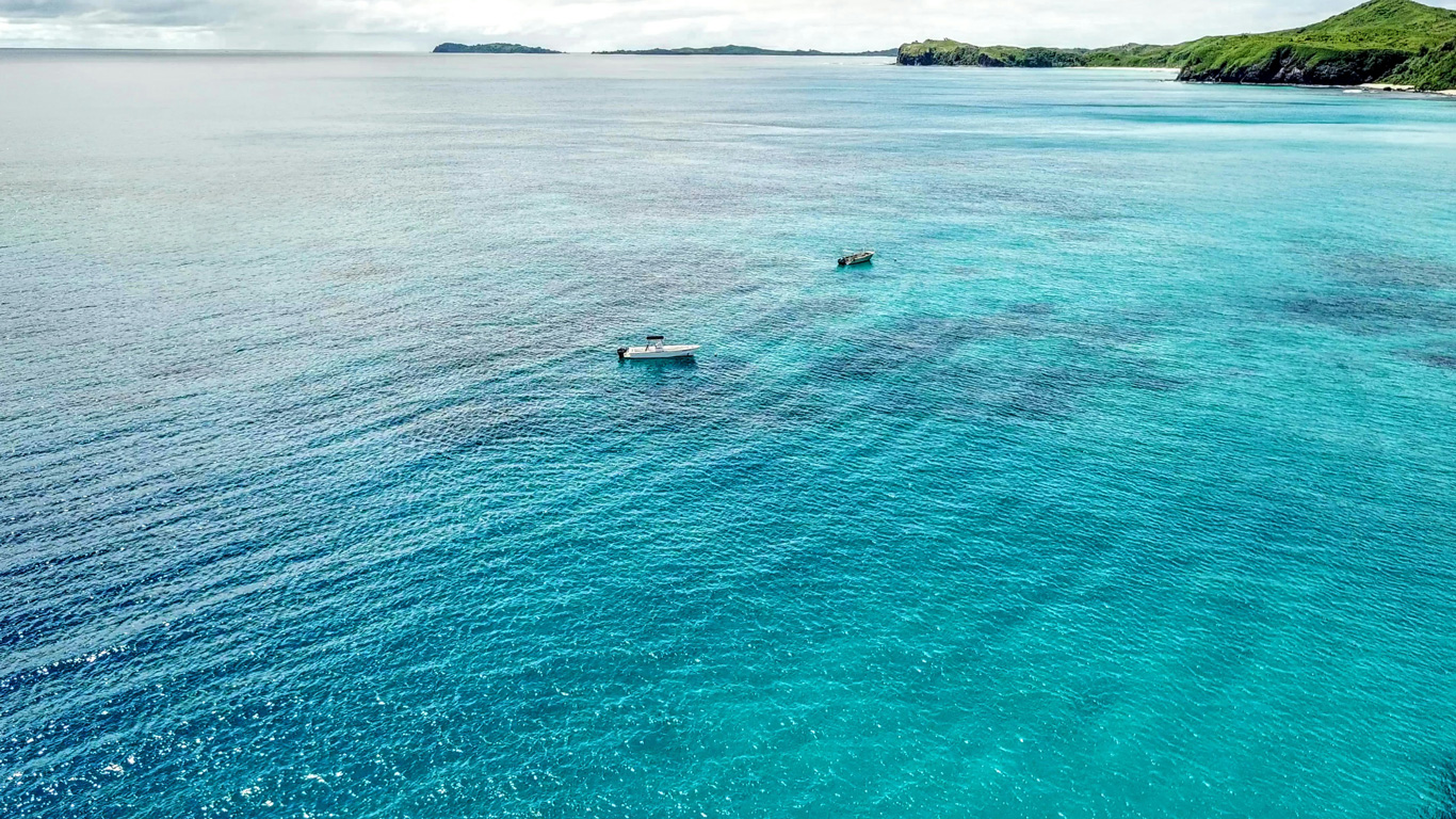 Two boats float on the crystal-clear turquoise waters near the Fiji Islands, with lush green coastline visible in the background. Gentle waves ripple across the ocean, reflecting the soft light of the sky. The peaceful and remote setting captures the essence of a tropical island paradise.