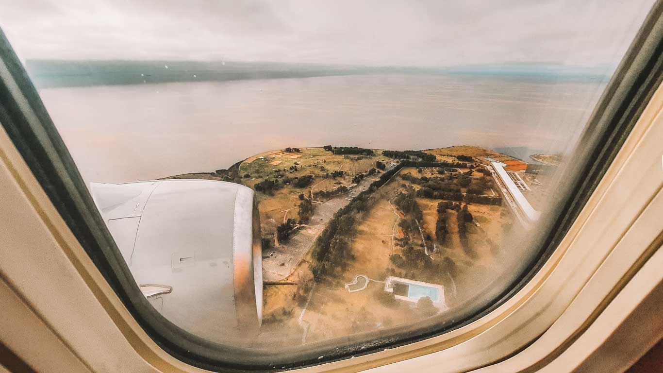 Aerial view from an airplane window showing a coastal landscape with open fields, a swimming pool, and a large building near the shoreline. The airplane engine is visible in the foreground, and the water stretches out under a hazy, overcast sky. The scene captures the approach to a destination, evoking the excitement of travel.