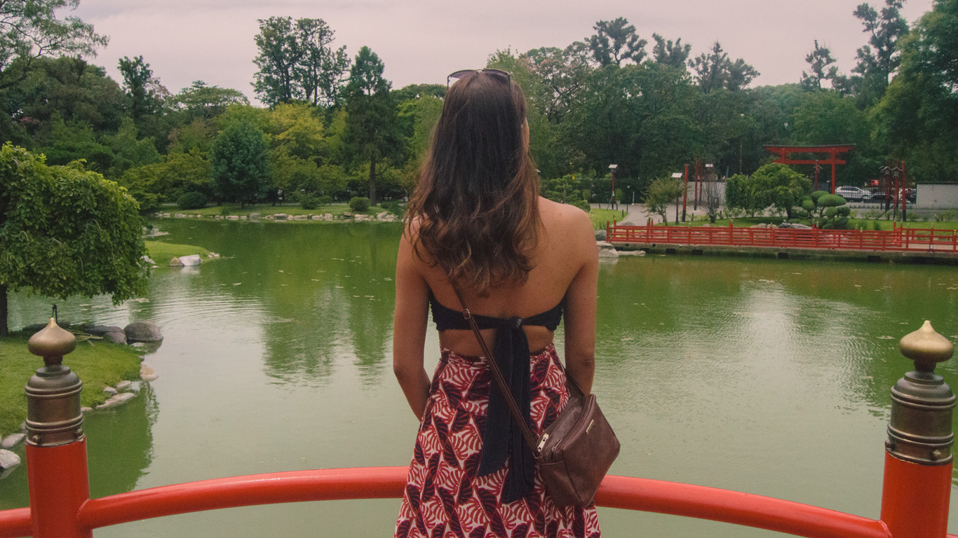 A woman with long brown hair stands on a red bridge overlooking a serene pond in the Japanese Garden of Buenos Aires. She wears a black bikini-style top, a patterned red skirt, and a brown crossbody bag, gazing at the lush greenery, stone-lined water, and traditional red torii gate in the distance. The peaceful garden setting reflects Japanese architectural elements, creating a tranquil and picturesque atmosphere.