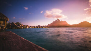 Scenic view of overwater bungalows in a tropical resort, possibly in Bora Bora, with a striking mountain peak in the background. The sunset casts a warm glow over the ocean and sky, blending shades of pink, orange, and purple. The calm waters reflect the light, creating a tranquil and luxurious ambiance.