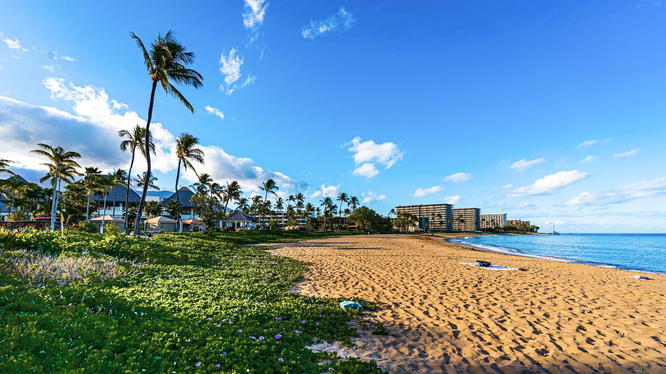 A scenic beach on Maui, Hawaii, with golden sand, lush greenery, and swaying palm trees under a clear blue sky. Luxury beachfront resorts and tropical villas line the shore, offering a picturesque island escape. Gentle waves lap against the coast, while a sailboat drifts in the distance, enhancing the tranquil ambiance.