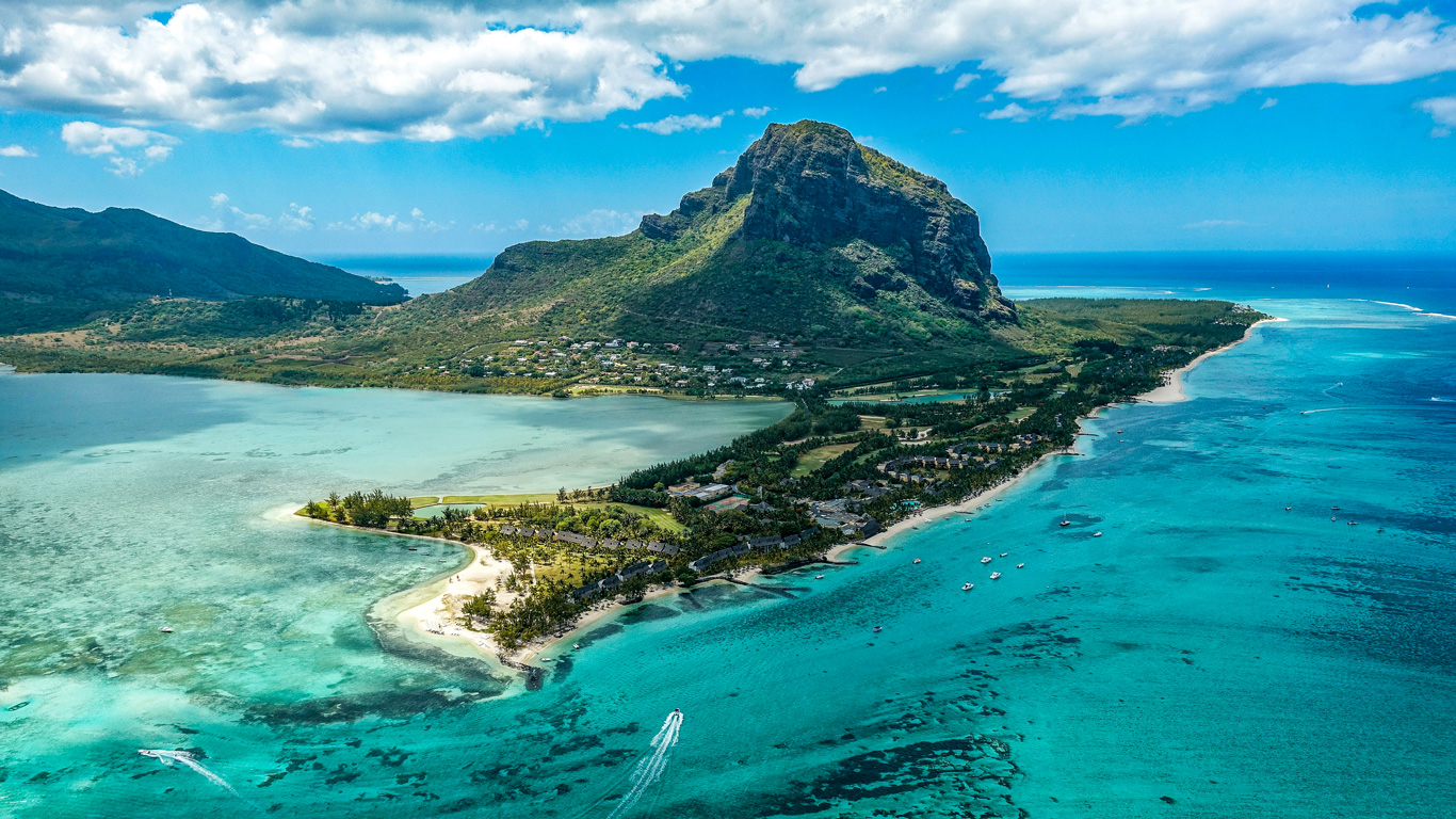 A breathtaking aerial view of Le Morne Brabant, a UNESCO World Heritage Site in Mauritius, surrounded by turquoise lagoons and lush greenery. The towering mountain dominates the landscape, with white sandy beaches and luxurious resorts nestled along the coastline. Boats glide across the crystal-clear waters, adding to the tropical paradise atmosphere.