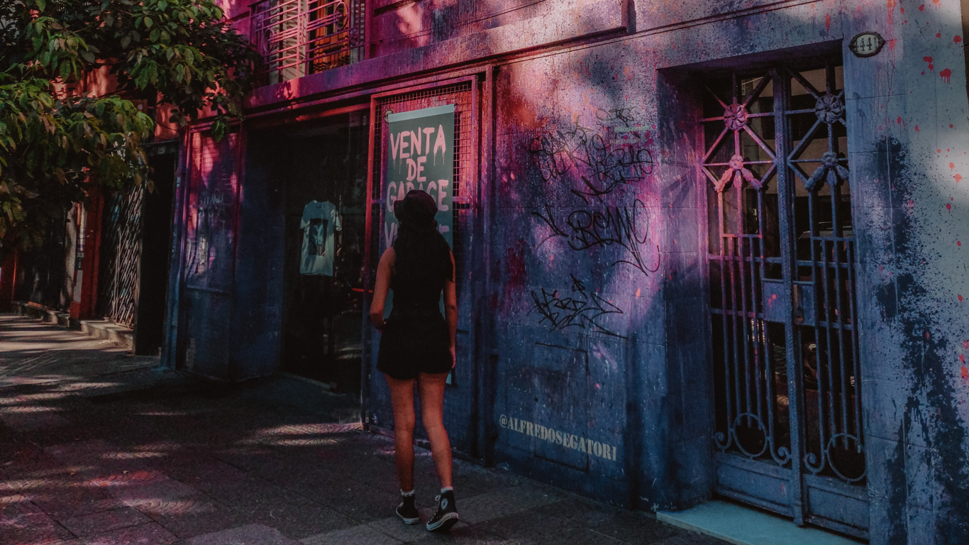 A woman dressed in black, wearing a hat and sneakers, stands in front of a graffiti-covered storefront in Palermo, Buenos Aires. The building's deep blue and purple tones are splattered with red paint, reflecting the edgy, artistic vibe of the neighborhood. A sign on the shop window reads 'Venta de Garage' (Garage Sale), adding to the urban, alternative atmosphere of the scene.