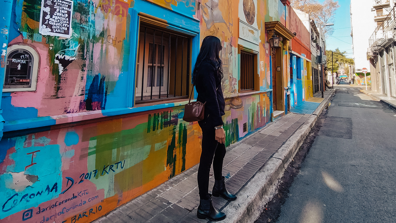 A woman dressed in black with long dark hair walks along Calle Russel in Palermo, Buenos Aires, admiring a building covered in vibrant, abstract street art. The walls are painted in bold shades of blue, orange, pink, and green, featuring graffiti, murals, and artistic lettering. The narrow street, lined with both colorful and neutral-toned buildings, exudes a bohemian and creative atmosphere.