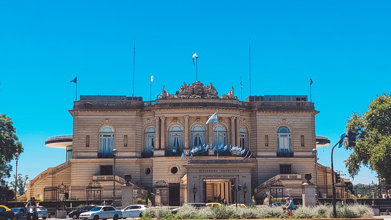 Front view of the Hipódromo Argentino de Palermo, a grand neoclassical horse racing venue in Buenos Aires, Argentina. The historic building features elegant arched windows, intricate sculptures on the rooftop, and multiple Argentine flags decorating the entrance. A busy street with cars, a motorcyclist, and a cyclist passes in front, adding to the vibrant city atmosphere.