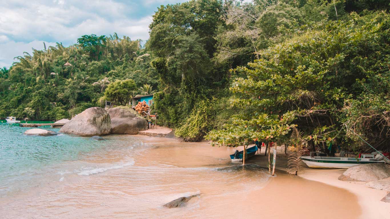 A small, secluded beach in Paraty, Rio de Janeiro, Brazil, with golden sand, turquoise waters, and lush green vegetation. Large smooth boulders sit at the shoreline, and a few colorful boats are moored under the shade of the trees. A blue house peeks out from the dense jungle, adding a charming, remote feel to the tropical setting.