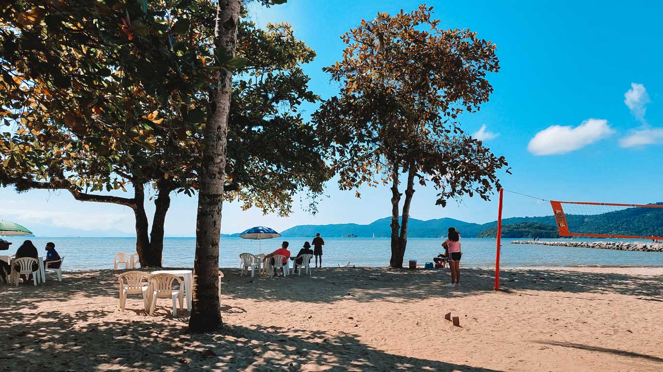 A serene beach in Paraty, Brazil, with people relaxing under umbrellas and sitting at white plastic tables in the sand. Trees provide shade in the foreground, while a volleyball net stands near the water’s edge. The calm ocean, distant islands, and a bright blue sky create a peaceful tropical atmosphere.