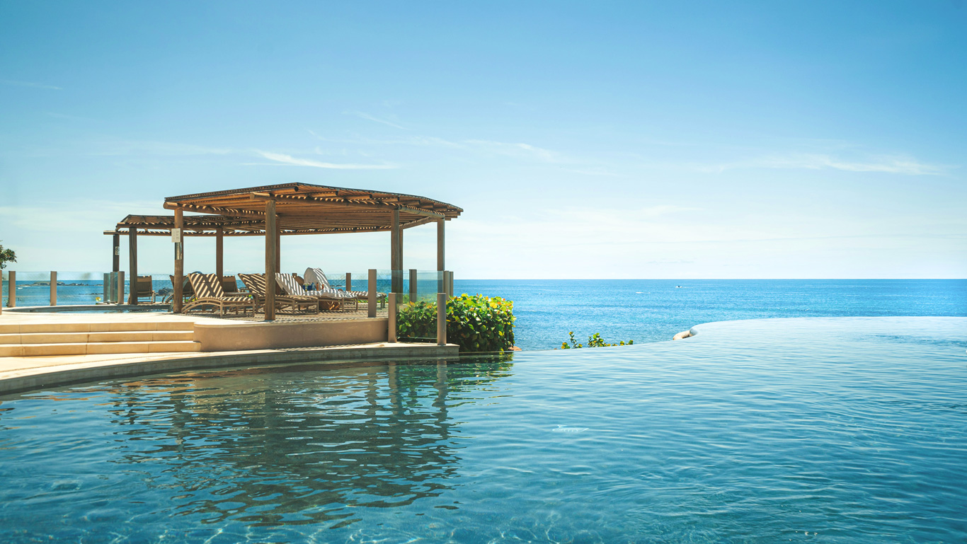 Serene oceanfront infinity pool at a luxury resort in Punta Mita, Mexico, seamlessly blending into the blue horizon. A shaded wooden pergola with lounge chairs offers a perfect relaxation spot beside the tranquil water. The clear sky and gentle ocean waves in the background enhance the peaceful, tropical ambiance.