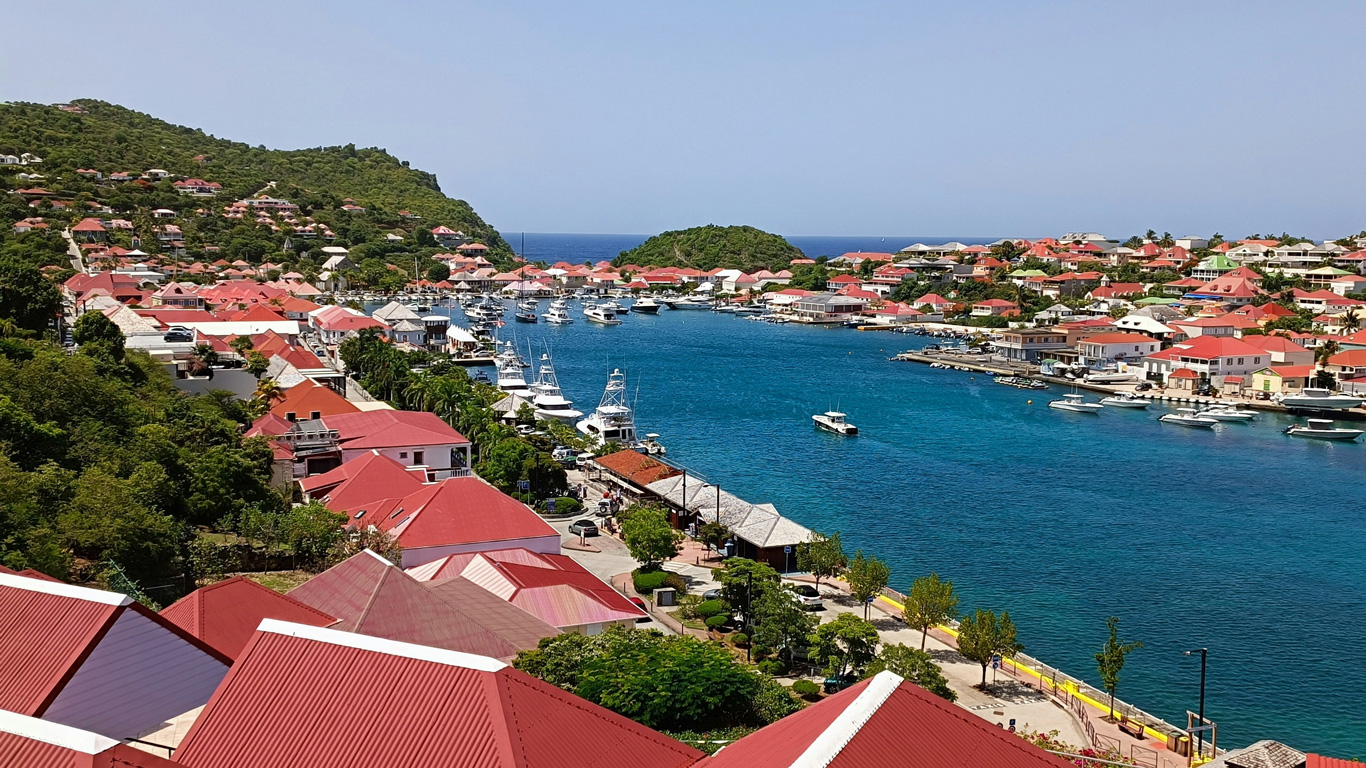 A picturesque view of Gustavia, the capital of Saint Barthélemy, featuring red-roofed buildings nestled along a turquoise harbor. Luxury yachts and boats are docked in the marina, surrounded by lush green hills. The vibrant waterfront, charming architecture, and clear blue sky highlight the island’s upscale Caribbean charm.