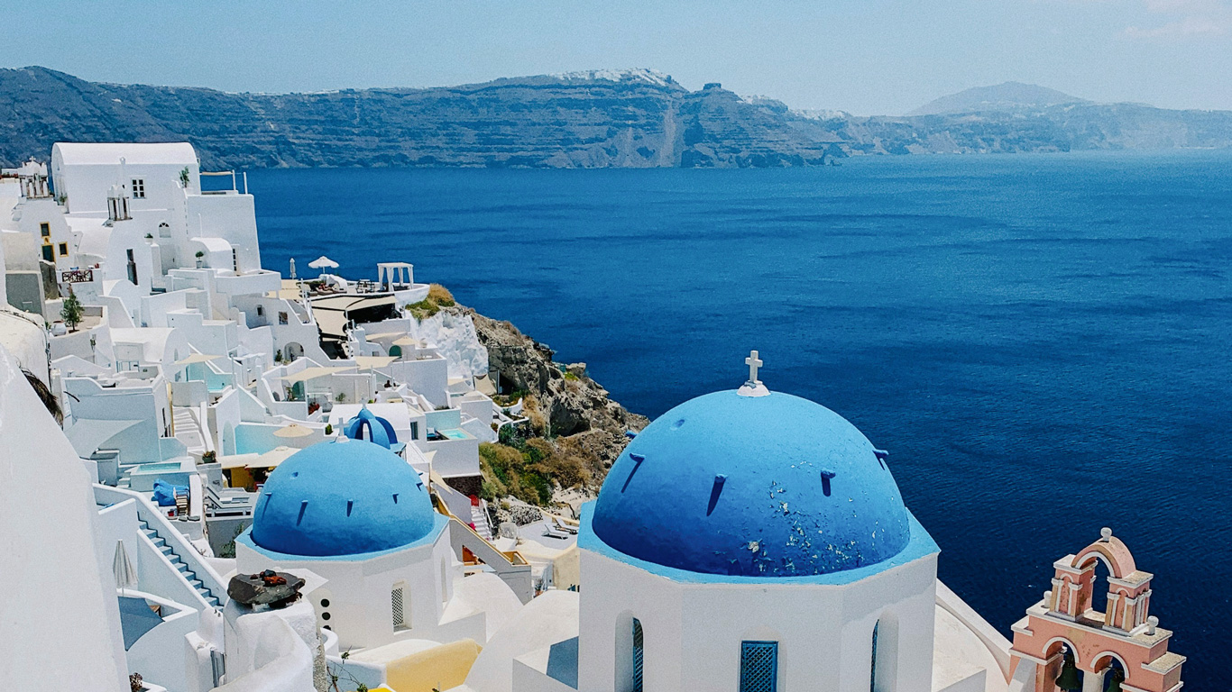 A stunning view of Santorini, Greece, featuring the iconic blue-domed churches and whitewashed buildings overlooking the deep blue Aegean Sea. The rugged cliffs and distant islands create a breathtaking backdrop. Sunlight casts a warm glow over the picturesque Cycladic architecture, highlighting the charm of this famous Mediterranean destination.