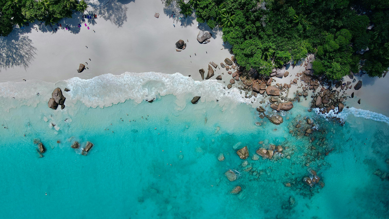 A stunning aerial view of a tropical beach in Seychelles, showcasing soft white sand, turquoise waters, and scattered granite boulders along the shoreline. Waves gently lap against the shore, while lush green vegetation frames the scene. Swimmers can be seen enjoying the clear water, adding to the serene and picturesque atmosphere.