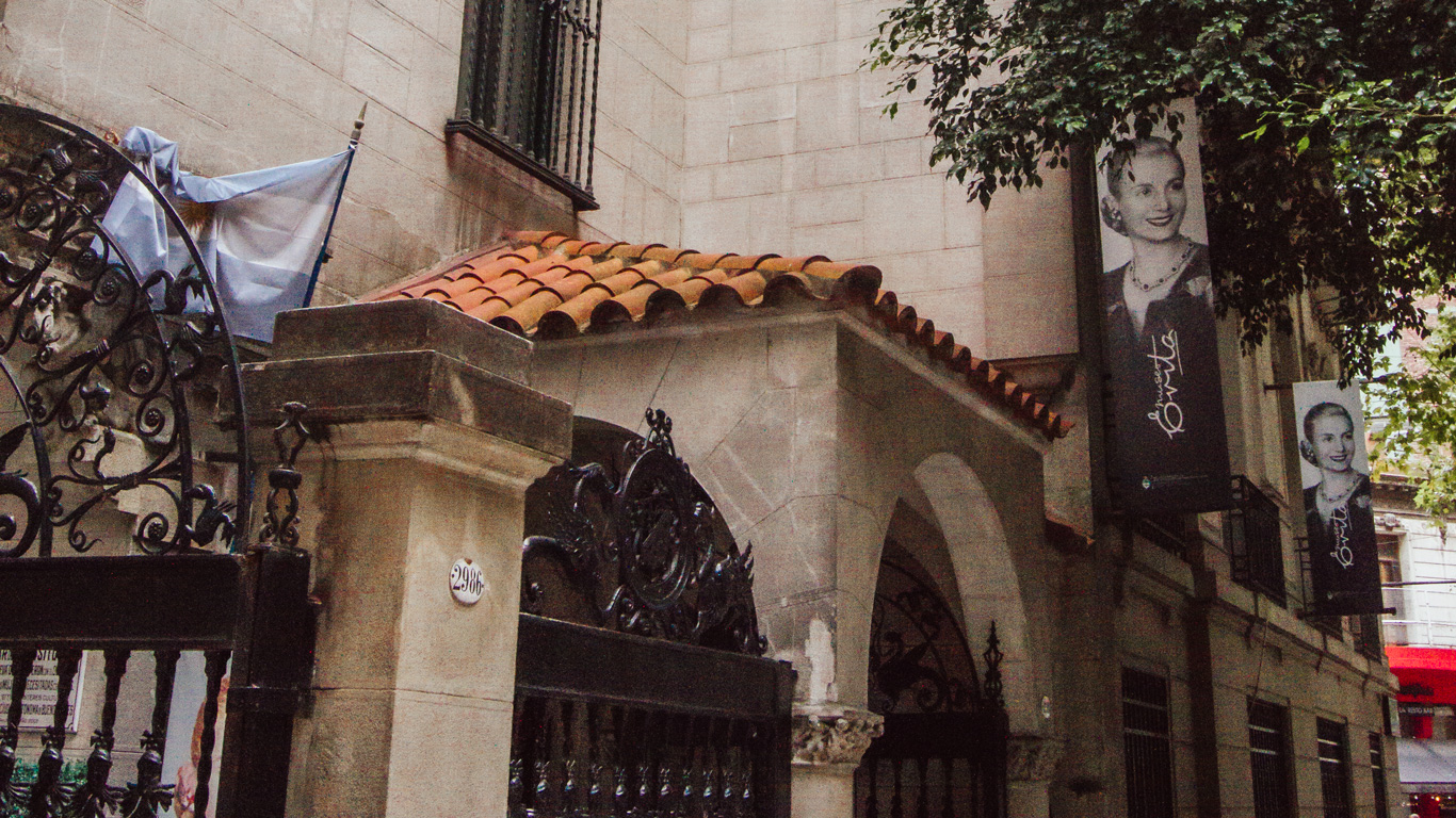 The entrance to the Evita Museum in Buenos Aires, Argentina, featuring an ornate black wrought iron gate and a stone façade with a terracotta-tiled roof. Above the entrance, banners display black-and-white portraits of Eva Perón, smiling and elegantly dressed. An Argentine flag hangs beside the entrance, adding to the historical and cultural significance of the site.