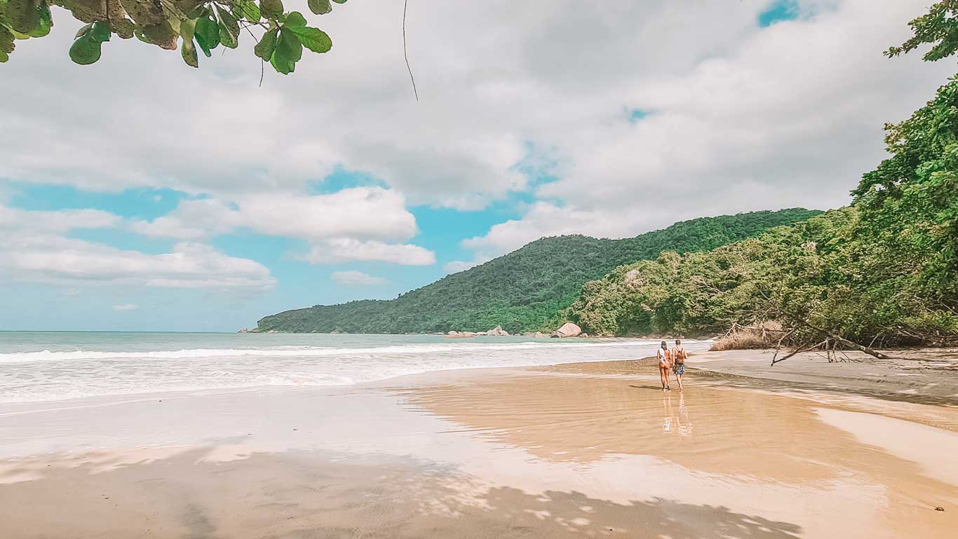 A secluded tropical beach in Trindade, Rio de Janeiro, with soft golden sand, gentle waves, and lush green hills in the background. Two people walk along the shoreline, leaving footprints in the wet sand as the ocean glistens under a partly cloudy sky. Overhanging tree branches frame the peaceful coastal scene.