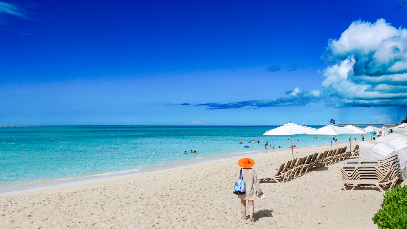 A woman in an orange sun hat walks along the pristine white sands of a beach in Turks and Caicos, with crystal-clear turquoise waters stretching to the horizon. Rows of lounge chairs and umbrellas line the shore, while swimmers enjoy the calm sea. The deep blue sky is dotted with fluffy clouds, creating a perfect tropical getaway scene.