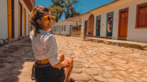 A woman wearing orange sunglasses, a white shirt, and black shorts sits on a cobblestone street in Paraty, Brazil, smiling under the sun. Behind her, colonial-style buildings with colorful doors and windows line the street, with hanging textiles and hats displayed outside. The warm, golden light enhances the historic charm of the scene.