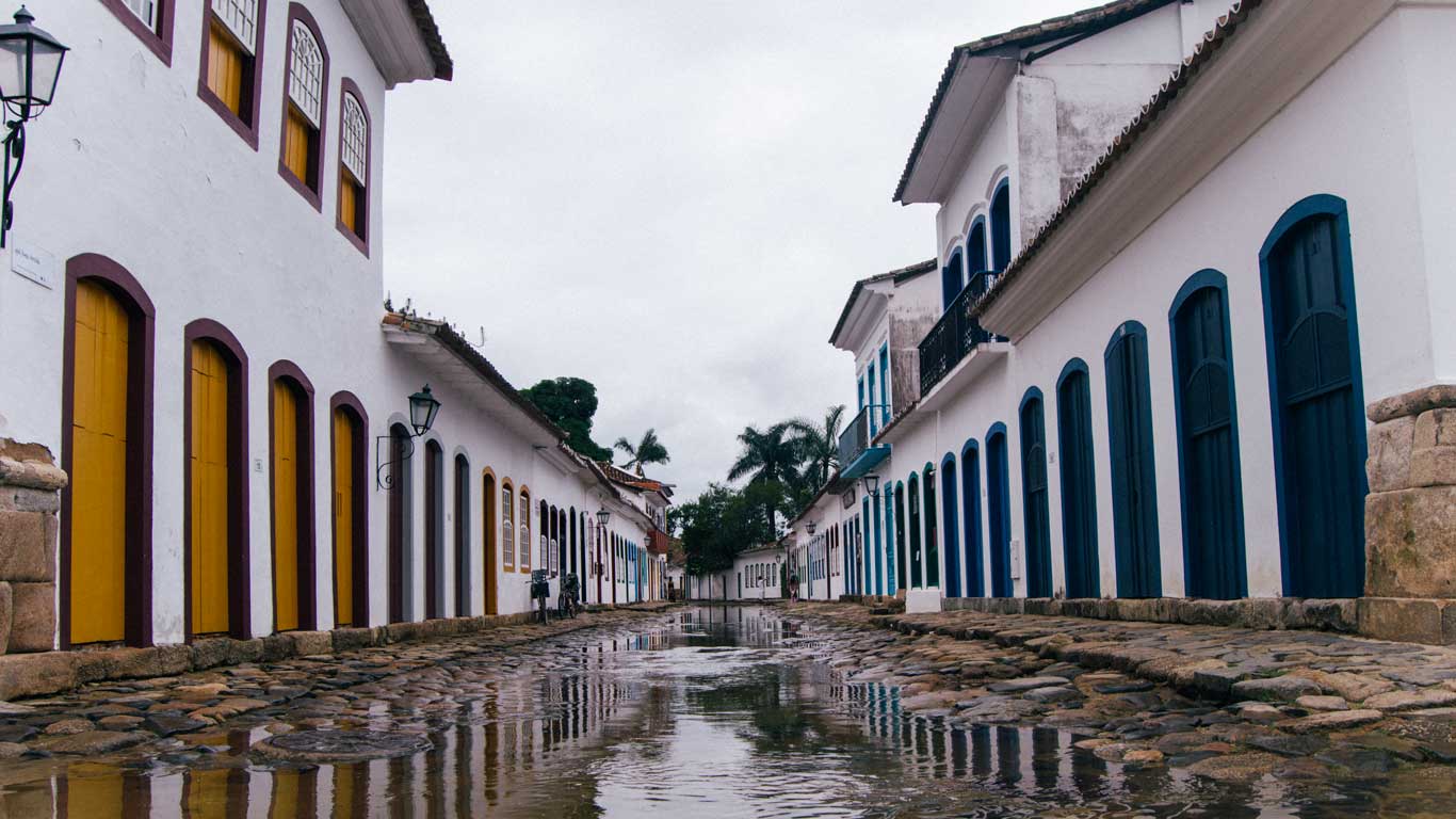 A flooded cobblestone street in Paraty, Brazil, reflecting the white colonial-style buildings with colorful doors and windows. The overcast sky and puddles add a moody, atmospheric feel to the historic town. Palm trees and vintage-style lanterns line the quiet street, emphasizing Paraty’s old-world charm.