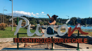 A woman in a blue striped dress and sunglasses poses playfully behind a large 'Pucón' sign that reads 'El Centro del Sur de Chile.' She points towards the snow-capped Villarrica Volcano in the background, with a scenic lakeside marina, boats, and modern buildings adding to the vibrant atmosphere. The bright blue sky and lush greenery enhance the beauty of this popular Chilean destination