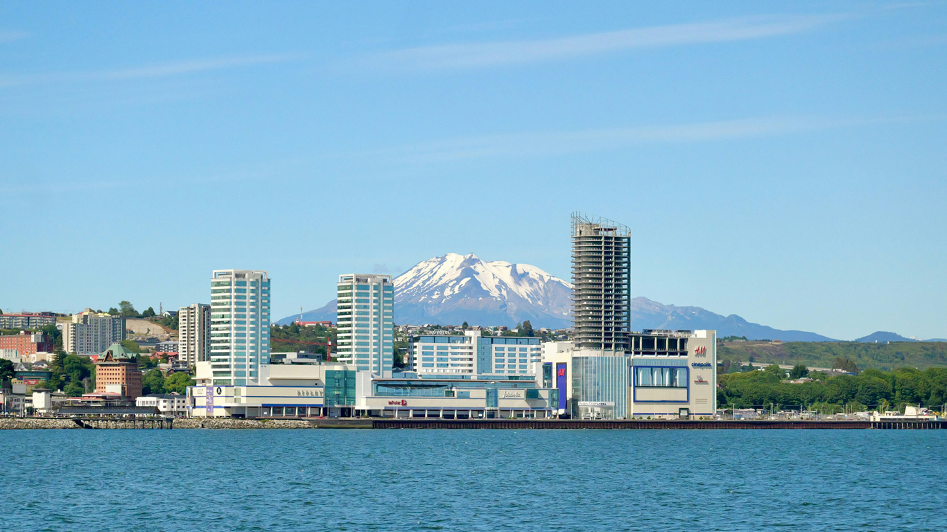 Scenic waterfront view of Puerto Montt, Chile, featuring modern high-rise buildings, a shopping mall, and a partially constructed tower along the coast. The calm blue waters in the foreground contrast with the snow-capped peaks of a majestic volcano in the background. The city skyline blends urban development with the region’s stunning natural landscape.