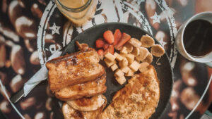 A hearty breakfast served on a black plate with golden French toast drizzled in syrup, a herb omelette, and fresh-cut fruit including strawberries, bananas, and diced melon, accompanied by a cup of black coffee and a glass of iced latte on a table with coffee bean-themed artwork.