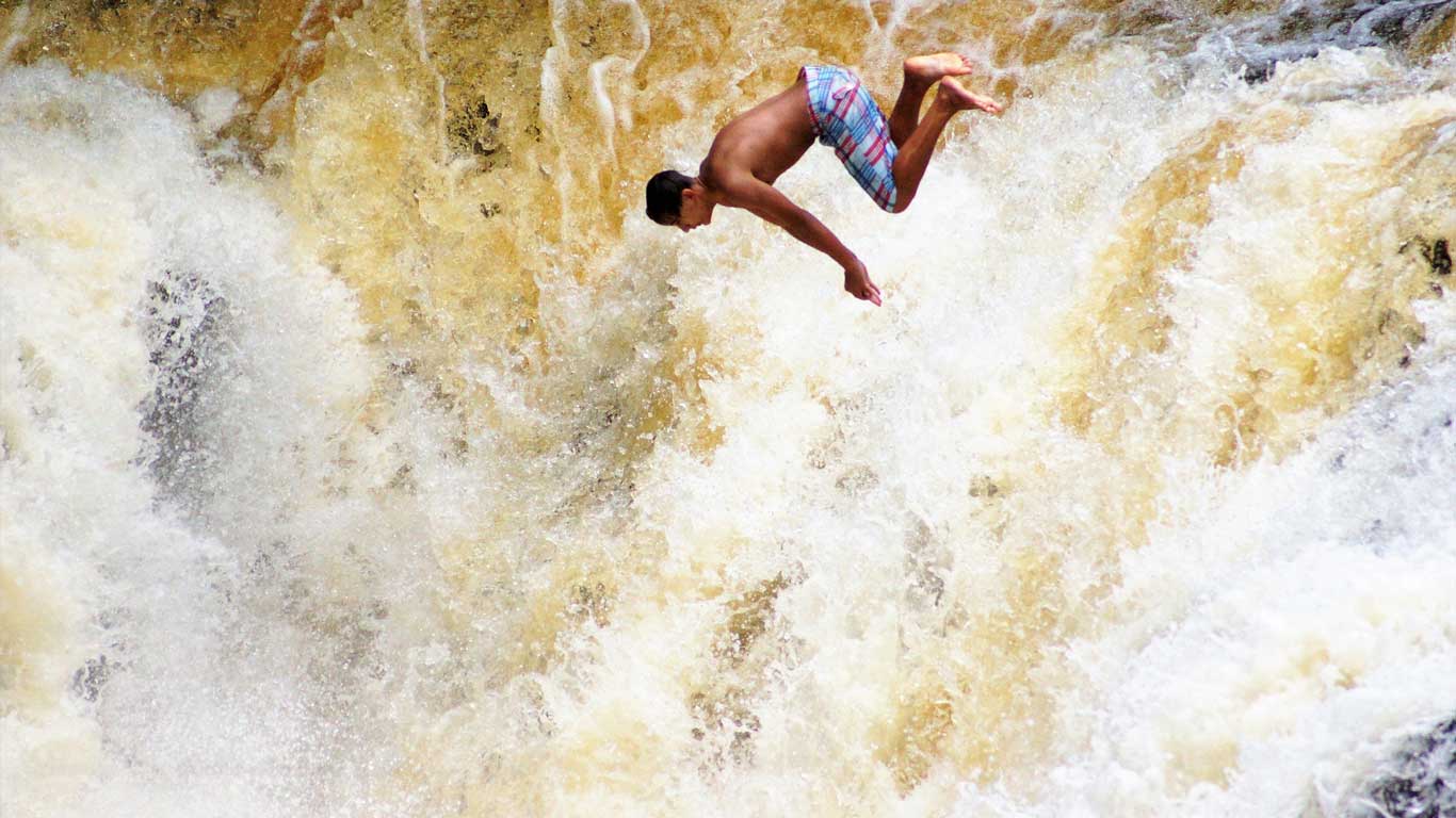 A young man in colorful swim trunks is captured mid-air as he performs a daring backflip into a rushing waterfall. The powerful, foamy water cascades down the rocky surface, creating a dramatic and adventurous scene. This image captures the thrill of waterfall diving, a popular activity in Brotas, São Paulo, known for its eco-tourism and extreme sports.