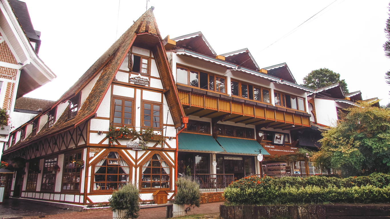 A European-style building with a steeply pitched roof, wooden beams, and white walls, resembling traditional Swiss or German architecture. The building houses the 'Matterhorn' restaurant and a few other businesses, with green awnings, wooden balconies, and flower boxes adding to its charm. The scene is set in Campos do Jordão, São Paulo, a popular mountain town known for its alpine aesthetics and cozy ambiance.