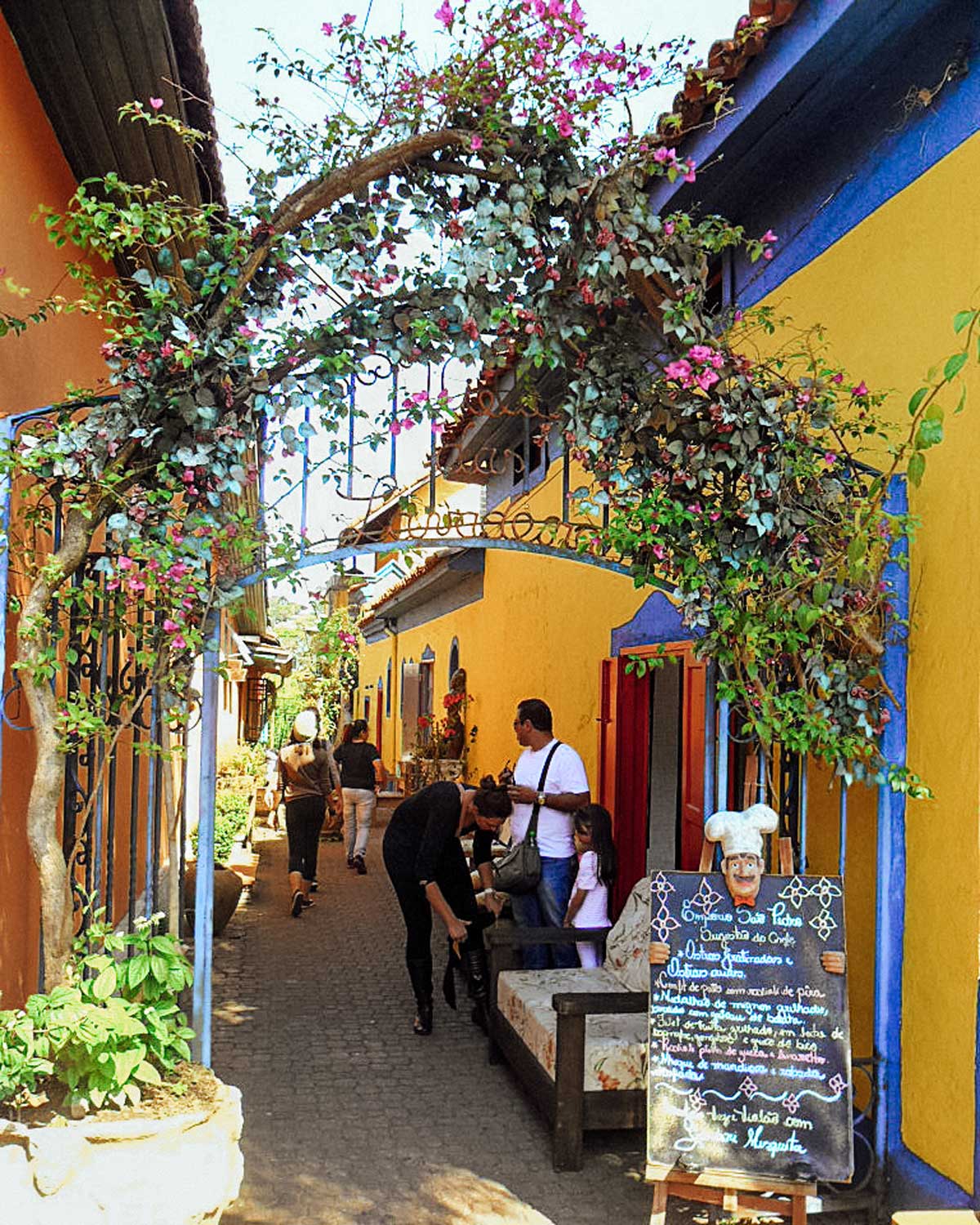 A charming cobblestone alleyway in Embu das Artes, Brazil, lined with colorful colonial-style buildings painted in warm yellow and blue tones. A decorative arch covered in pink bougainvillea flowers frames the entrance, leading to a cozy café with a chalkboard menu beside a small chef figurine. People walk through the picturesque street, exploring the vibrant artisan shops and cafés that define this artistic town.