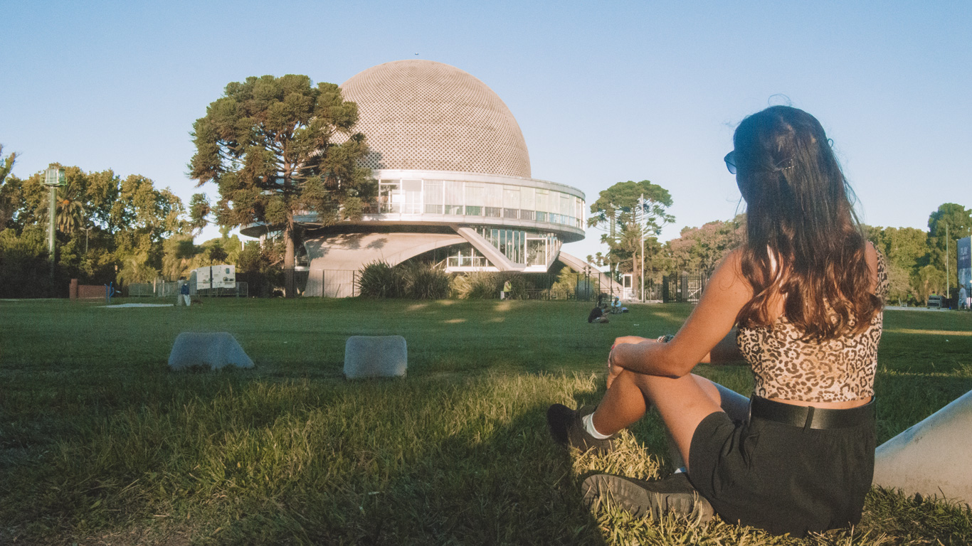 A woman with long brown hair sits on the grass in front of the Galileo Galilei Planetarium in Buenos Aires, Argentina. She wears a leopard-print top, black shorts, and sneakers, gazing at the futuristic dome-shaped building with its modern architecture and glass windows. The surrounding park is bathed in warm sunlight, with scattered trees and a few people relaxing in the open green space.