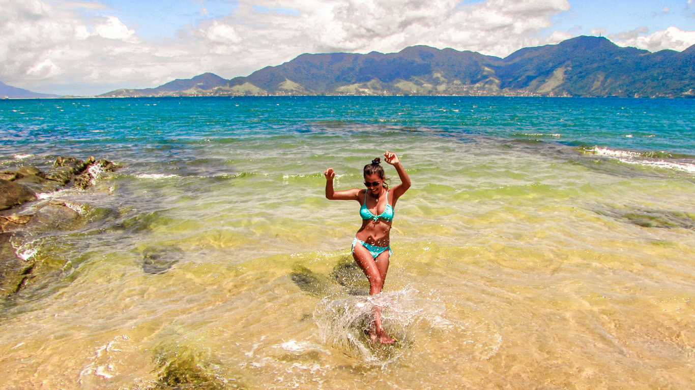 A woman in a turquoise bikini splashes in the shallow waters of a tropical beach in Ilhabela, São Paulo, Brazil. The crystal-clear sea transitions from light green near the shore to deep blue further out, with a backdrop of lush green mountains under a partly cloudy sky. She smiles joyfully, embracing the vibrant and refreshing beach setting.