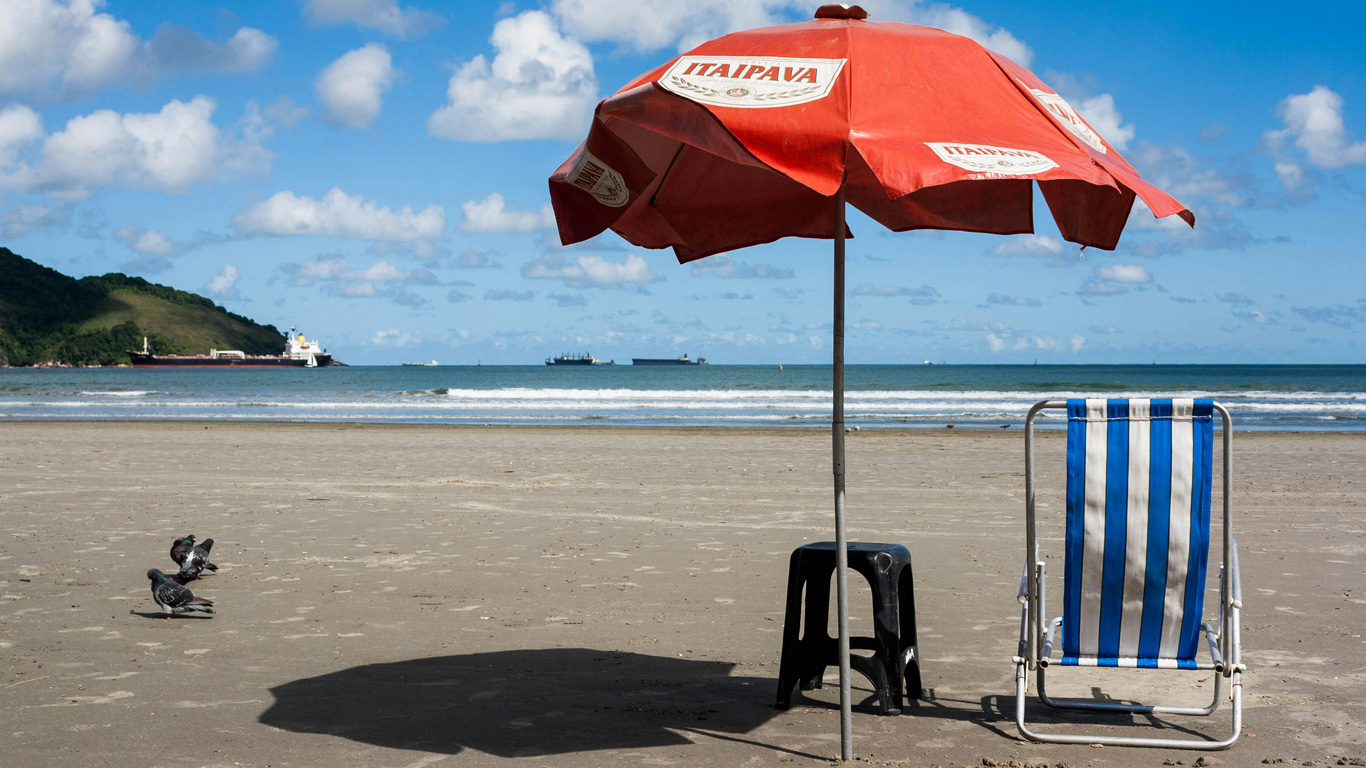 A peaceful beach scene in Santos, Brazil, featuring a red Itaipava-branded beach umbrella shading a plastic stool and a blue-and-white striped chair. A few pigeons peck at the sand nearby, while cargo ships float on the horizon against the deep blue ocean. The background showcases a green hill and a bright blue sky dotted with fluffy white clouds.