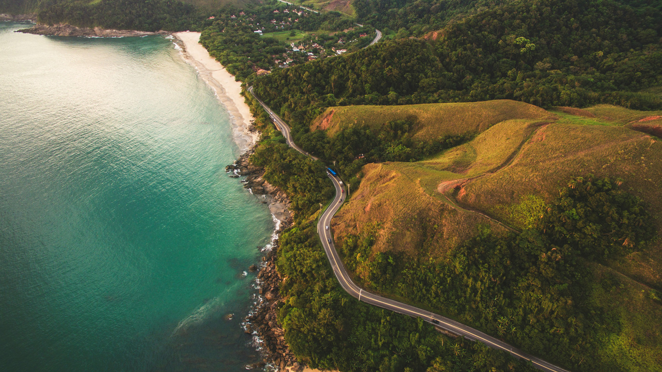 A breathtaking aerial view of São Sebastião, Brazil, showcasing a winding coastal road hugging lush green hills. The turquoise ocean meets a secluded sandy beach, while a small village is nestled among the trees in the background. The golden light casts a warm glow over the landscape, highlighting the natural beauty of this tropical paradise.