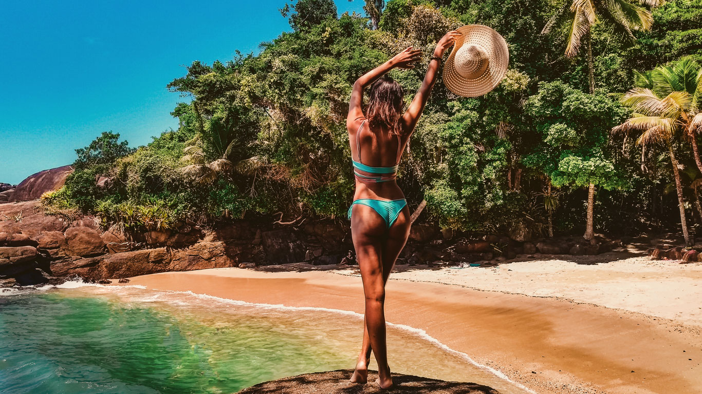 A woman in a turquoise bikini stands on a rocky ledge overlooking a secluded tropical beach in Ubatuba, São Paulo, Brazil. She raises a wide-brimmed sun hat in one hand, facing the turquoise water and golden sand, framed by lush green palm trees and dense jungle. The bright blue sky and untouched shoreline create a dreamy, exotic paradise feel