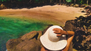 A woman wearing a wide-brimmed white sun hat sits on a rocky outcrop, overlooking a secluded beach in Ubatuba, São Paulo, Brazil. The turquoise-green water gently meets the golden sand, where a few people relax under the shade of tropical trees. Sunlight filters through the leaves, casting dappled shadows on the rocks, creating a tranquil and inviting atmosphere.