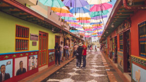 A vibrant cobblestone street in Guatapé, Colombia, lined with brightly painted buildings and colorful murals, with rows of suspended umbrellas creating a festive overhead canopy. People are strolling, chatting, and taking photos in the lively atmosphere.