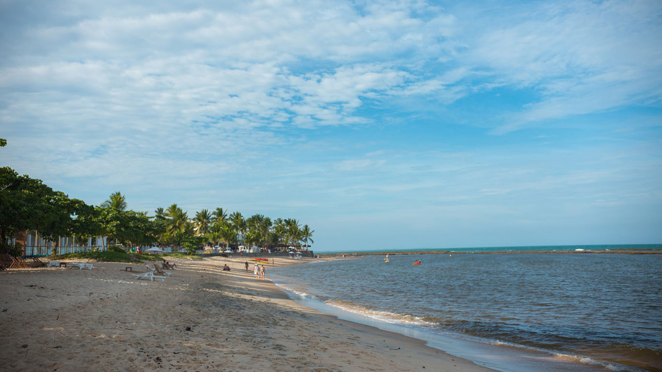apaga-fogo-beach Apaga Fogo Beach stretches along calm, shallow waters with a peaceful vibe and fewer visitors. Palm trees and a few buildings are visible in the distance, with gentle waves meeting the sandy shore beneath a partly cloudy sky.