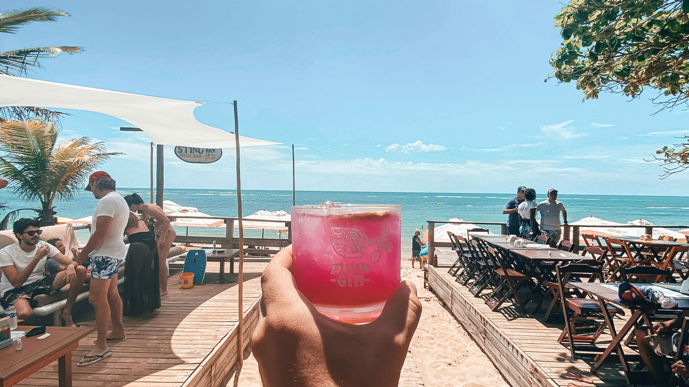 aracaípe-beach-arraial-ajuda A beachfront bar scene at Aracaípe Beach in Arraial d'Ajuda, with wooden decks, sun umbrellas, and a pink cocktail in the foreground—an inviting spot for anyone wondering where to stay in Arraial d'Ajuda for great drinks and ocean views.