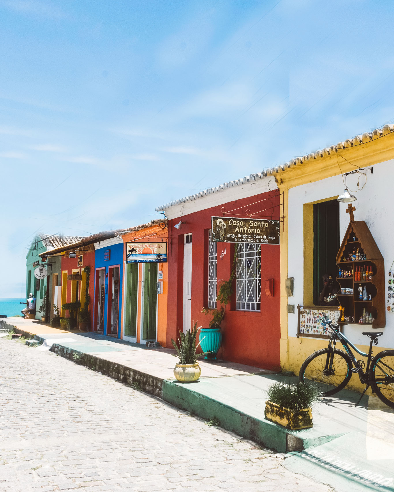 best-place-to-stay-arraial-dajuda A sunny street in Arraial d'Ajuda featuring vibrant buildings painted red, orange, green, and blue, with shops and signs visible. A potted plant, bicycle, and display of colorful ribbons and souvenirs decorate the entrance of a local store.