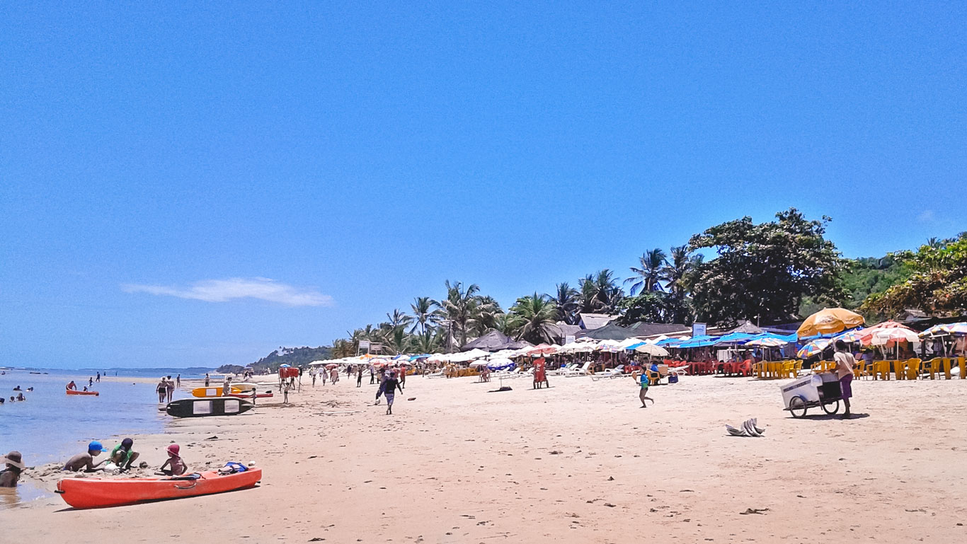 mucuge-beach-bahia Mucugê Beach in Bahia, Brazil, is bustling with activity as people enjoy the sandy shoreline and calm waters. Colorful umbrellas and beachside vendors line the shore, while children play near kayaks and canoes under a clear blue sky.