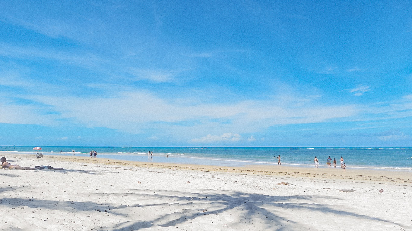 parracho-beach-arraial-dajuda Parracho Beach in Arraial d'Ajuda features soft white sand, calm turquoise water, and scattered beachgoers under a wide blue sky—ideal for those searching where to stay in Arraial d'Ajuda near peaceful coastal beauty.