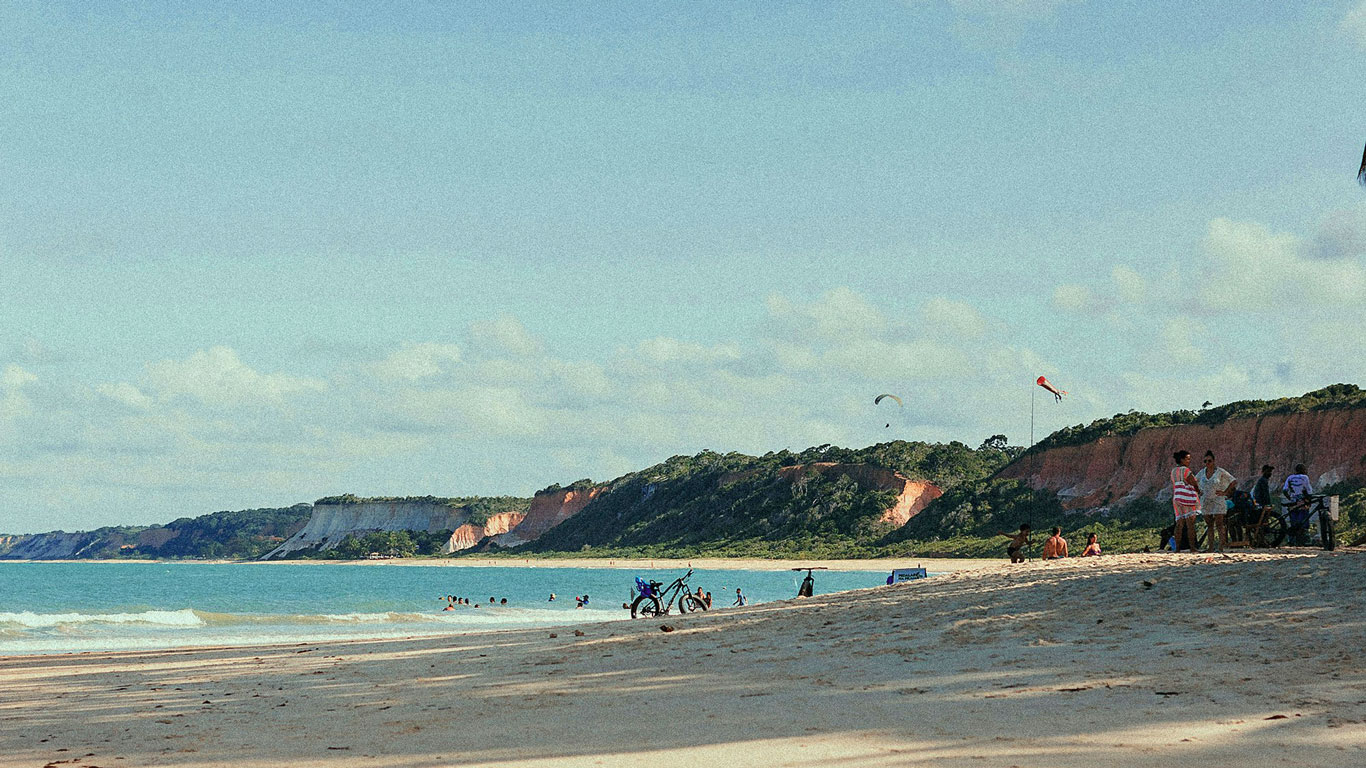 pitinga-beach-arraial-ajuda Pitinga Beach in Arraial d'Ajuda showcases dramatic red cliffs, lush greenery, and a sandy beach with swimmers and paragliders.