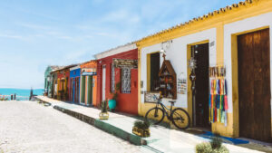 Colorful colonial-style storefronts line a cobblestone street in Arraial d'Ajuda, Bahia, with ocean views in the background. A black bicycle is parked outside a souvenir shop selling religious and handcrafted items, under a bright blue sky.