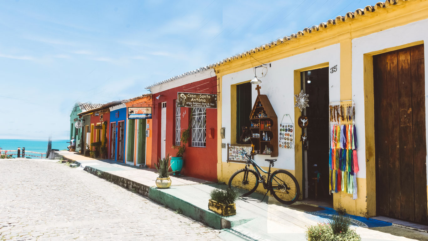 where-to-stay-in-arraial-dajuda-bahia Colorful colonial-style storefronts line a cobblestone street in Arraial d'Ajuda, Bahia, with ocean views in the background. A black bicycle is parked outside a souvenir shop selling religious and handcrafted items, under a bright blue sky.