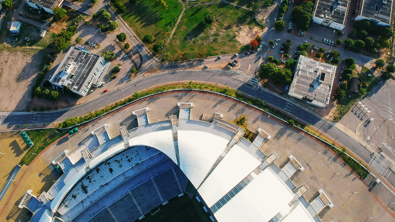 arena-das-dunas-natal Aerial view of Arena das Dunas stadium in Natal, Brazil, showing its white curved roof, surrounding roads, and nearby urban layout with green areas and buildings.
