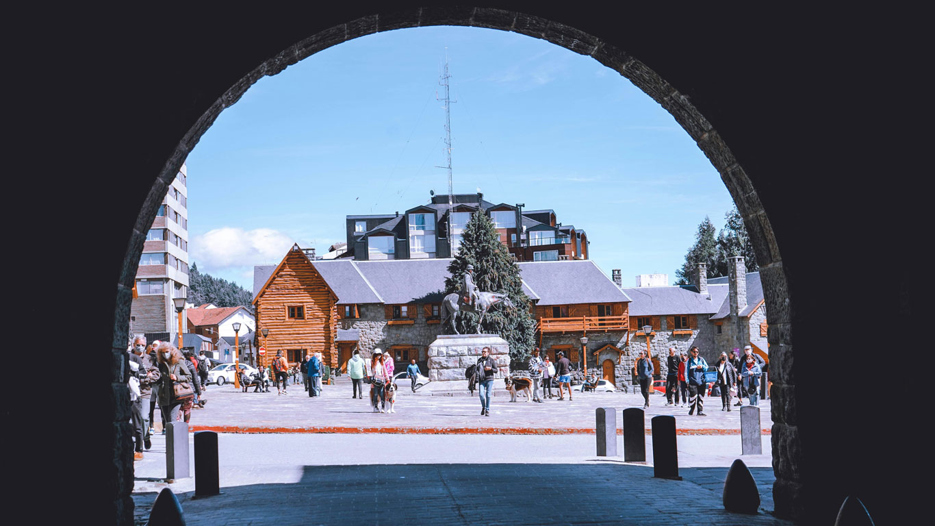 bariloche-in-the-winter A busy town square in Bariloche during winter, framed by a stone archway, with people walking near a statue, rustic alpine-style buildings, and bright blue skies overhead.