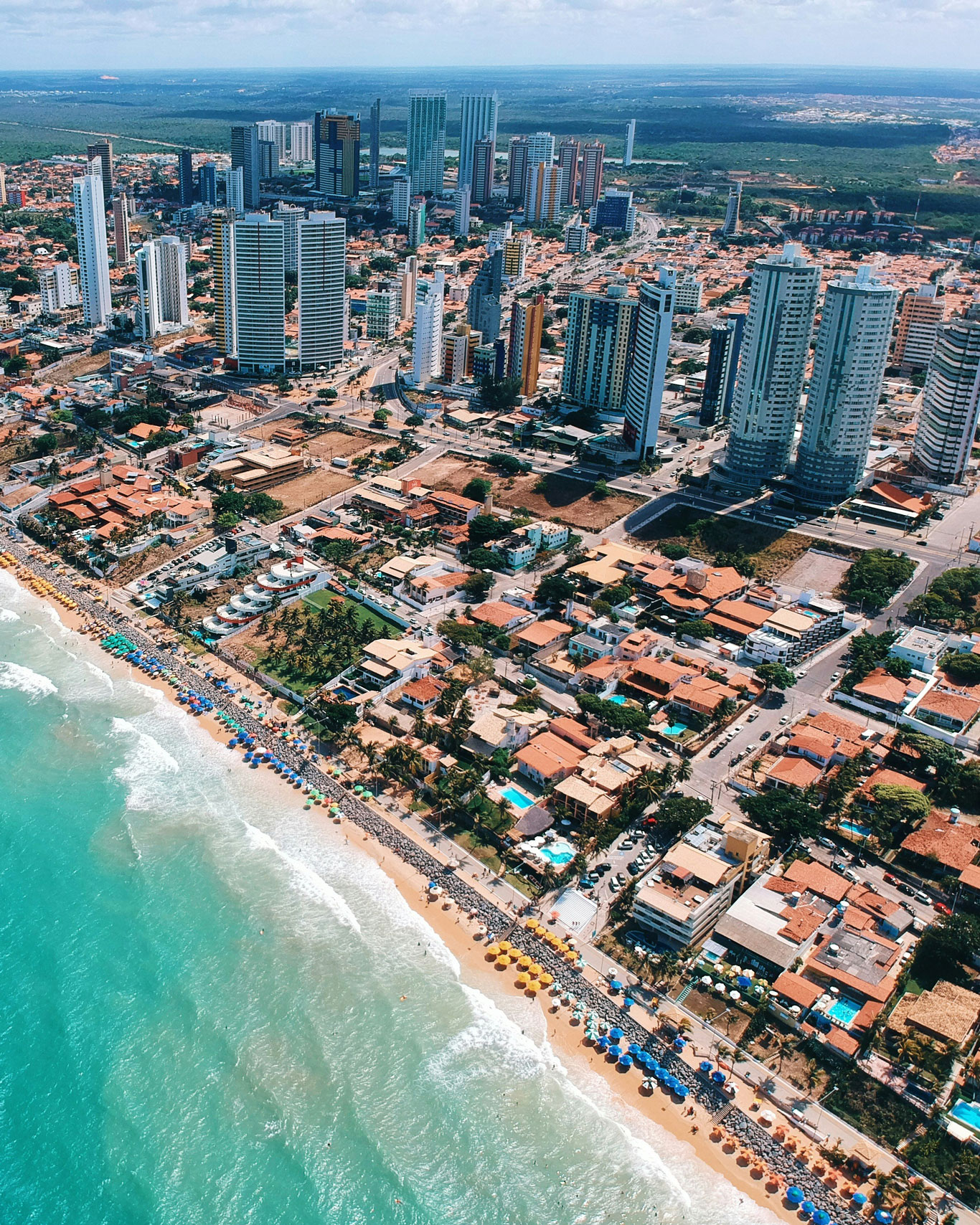 best-place-to-stay-in-natal Aerial view of a modern coastal cityscape in Natal, Brazil, with high-rise hotels and apartments close to the beach, and colorful umbrellas and beachgoers forming a lively strip along the water.