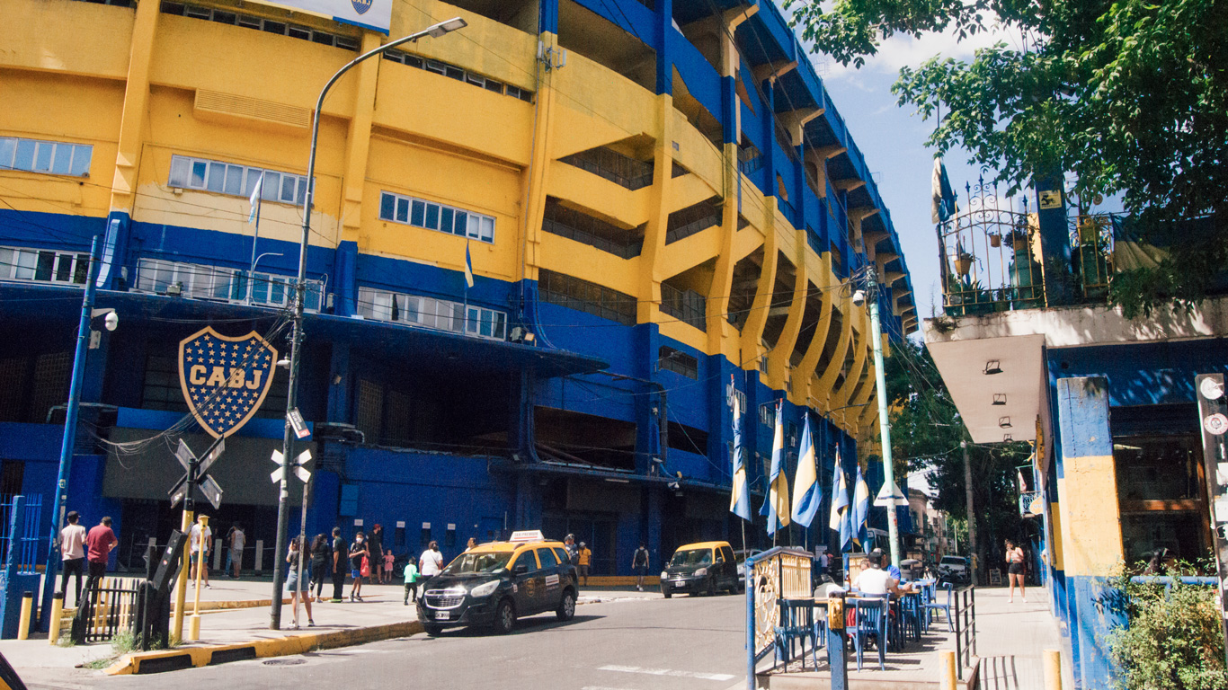 boca-juniors The vibrant yellow and blue exterior of La Bombonera stadium, home to Boca Juniors, with people and taxis around the entrance and flags flying proudly.