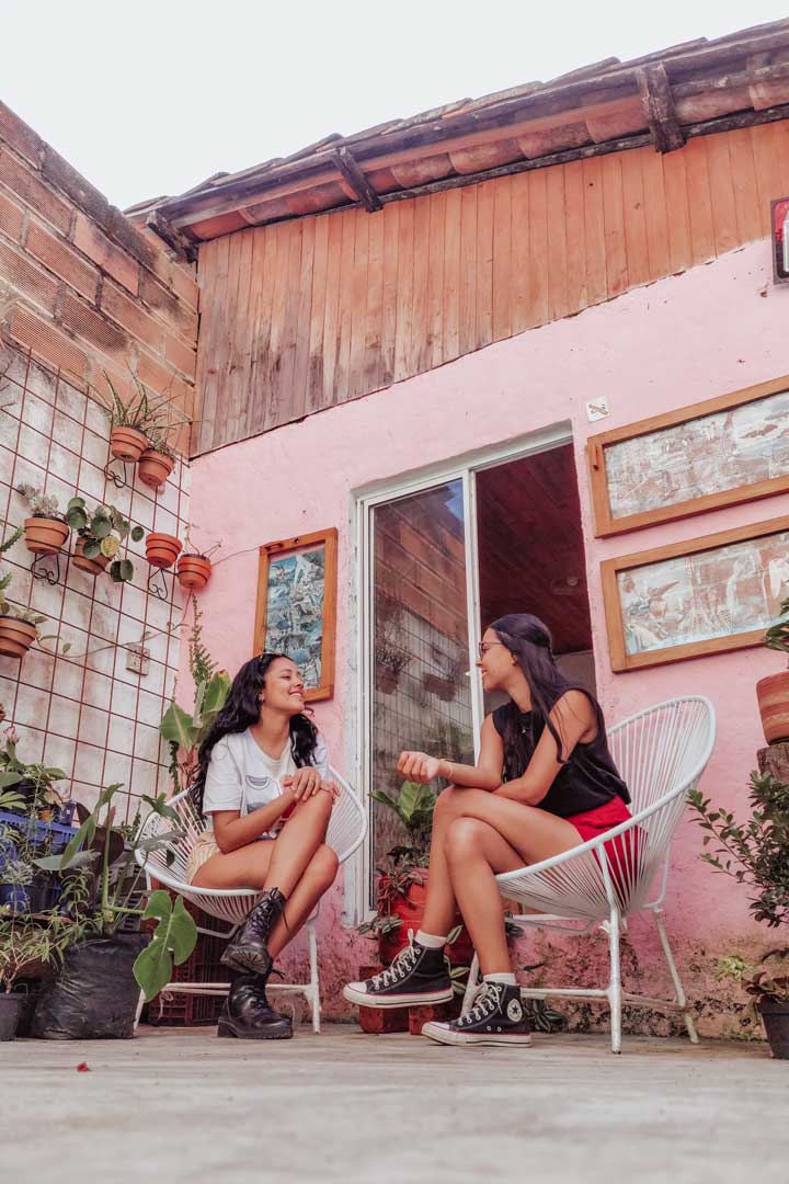 cafe-el-jardin-de-jardin Two women sit across from each other in white patio chairs, laughing and chatting in a cozy courtyard café in Jardín, Colombia, surrounded by potted plants and rustic decor.
