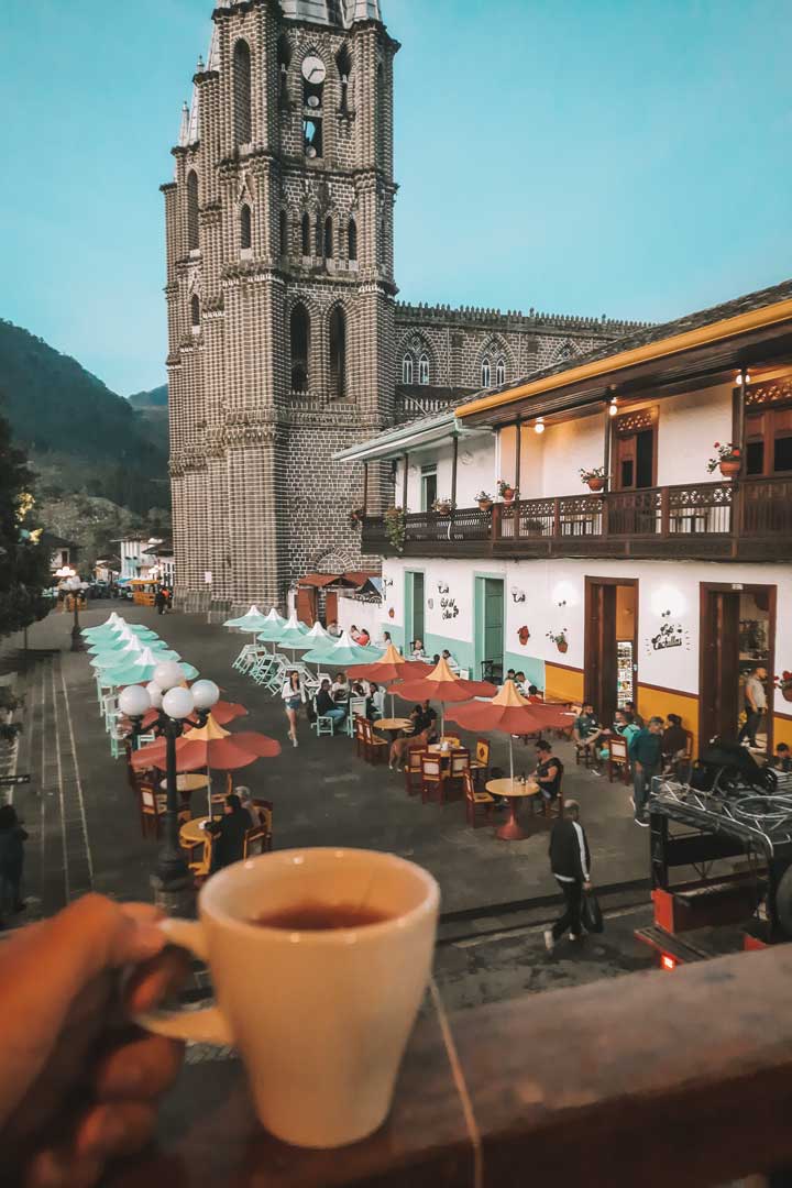 cafe-los-andes-jardin A hand holds a cup of coffee on a balcony overlooking a charming plaza in Jardín, Colombia, with colorful umbrellas and people dining below. In the background stands the illuminated Basilica of the Immaculate Conception, its brick facade glowing softly as evening sets in.