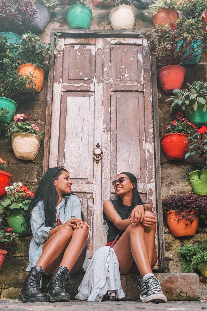 cafe-macanas-in-jardin Two women sit smiling on stone steps in front of a rustic wooden door, surrounded by a vibrant wall of colorful flower pots in Jardín, Colombia.